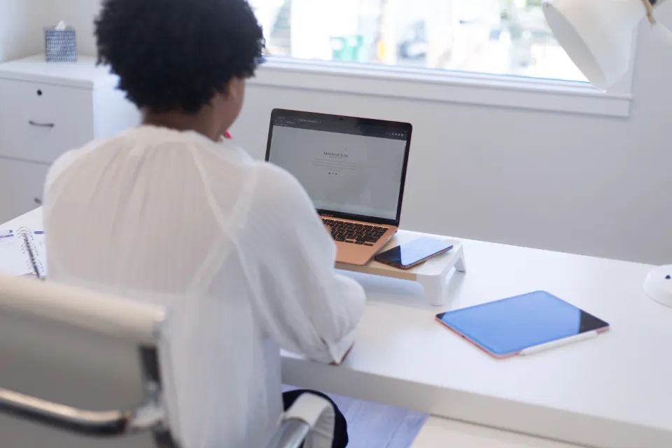A woman at a white desk working on a laptop, with a tablet beside her
