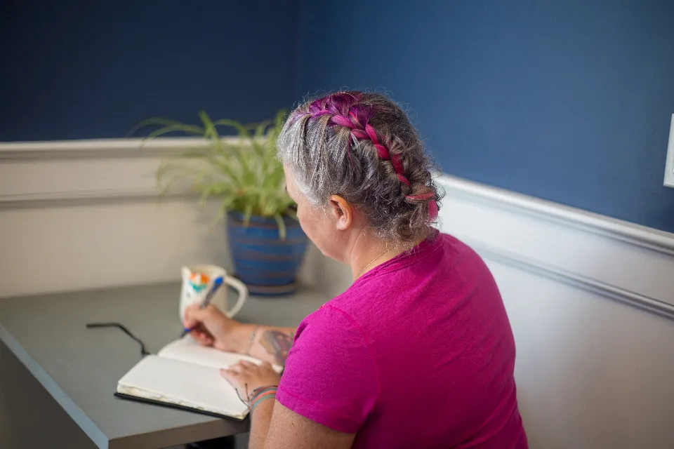 A woman with grey hair writing notes at a desk beside a window